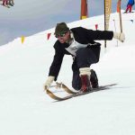 A telemark skier wearing vintage clothing and a hat bends low while skiing downhill on wooden skis in a snowy, mountainous area.