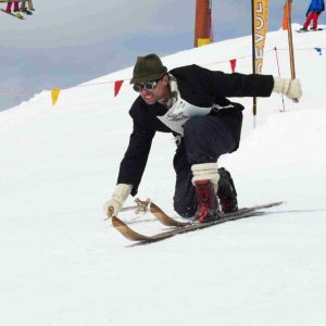 A telemark skier wearing vintage clothing and a hat bends low while skiing downhill on wooden skis in a snowy, mountainous area.