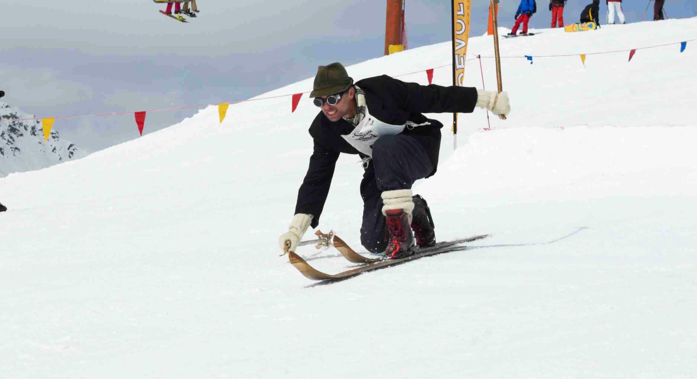 A telemark skier wearing vintage clothing and a hat bends low while skiing downhill on wooden skis in a snowy, mountainous area.