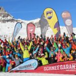 A large group of people in colorful ski gear pose on a snowy mountain, smiling and raising their arms. Behind them are promotional flags and a backdrop of snowy peaks under a clear blue sky.
